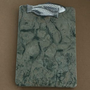 May include: A rectangular clipboard featuring a green and gray marbled stone surface. At the top, a silver-toned metal clip is shaped like two overlapping fish, providing a secure mechanism for holding papers in place on the flat stone board.