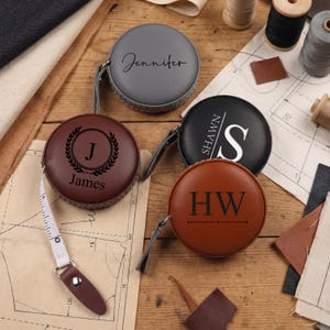 Peut inclure: Quatre rubans à mesurer rétractables ronds en similicuir gris, noir, brun foncé et havane. Chacun présente un texte ou des monogrammes personnalisés : Jennifer, James avec un J dans une couronne de laurier, Shawn avec un grand S et les initiales HW.