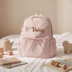Pode incluir: Uma mochila rosa claro com alça superior, um bolso frontal com detalhe de laço e texto bordado em marrom Veirty ao lado de um ícone de coração. A mochila tem um acabamento em tecido texturizado e bolsos laterais com fechos de correr.