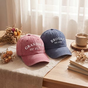Pode incluir: Um boné de beisebol rosa empoeirado com o texto branco GRANDMA EST 2027 e um boné azul-marinho com o texto branco BROTHE EST 2028. Ambos os bonés apresentam uma aba curva, uma coroa estruturada e um botão no topo, exibindo um design clássico.