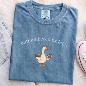 Könnte beinhalten: Ein blaues kurzärmeliges T-Shirt mit einer Grafik einer Gans, die eine Baseballkappe trägt und einen roten Spielzeuglaster sowie einen braunen Football hält. Der Text "outnumbered by boys" ist in Weiß über der Gans aufgedruckt.