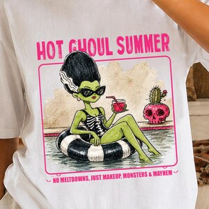 May include: A white t-shirt featuring a graphic of a green-skinned character with a tall black hairstyle, wearing sunglasses and a black swimsuit, floating in a pool on a black and white inner tube. The text reads HOT GHOUL SUMMER and NO MELTDOWNS, JUST MAKEUP, MONSTERS & MAYHEM.