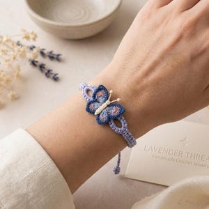 May include: A light purple crochet bracelet featuring a central butterfly motif. The butterfly has dark blue wings with pink and light blue embroidery details, and a cream-colored body with two small antennae. The band consists of linked loops.