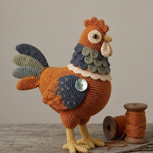 Puede incluir: Una figura de pollo tejida a ganchillo con cuerpo color naranja óxido, un ala azul oscuro con un botón de nácar brillante y una cola con plumas azules y verde salvia. El cuello tiene un plumaje superpuesto en forma de escamas azul y verde.
