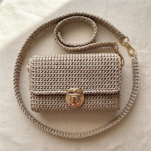 May include: A beige crochet crossbody bag featuring a rectangular body with a textured stitch pattern. The bag includes a gold-tone metal push-lock closure on the front flap and a matching braided crochet shoulder strap attached with gold-tone clips.