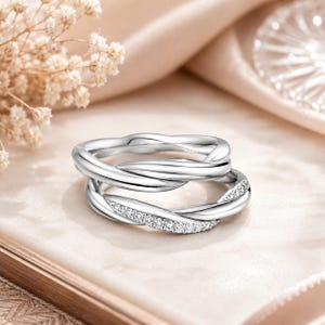 Peut inclure: Une bague argentée composée de deux anneaux entrelacés. L'anneau supérieur présente une finition lisse et polie, tandis que l'anneau inférieur est orné d'une rangée de petites pierres claires et scintillantes sur la surface torsadée.