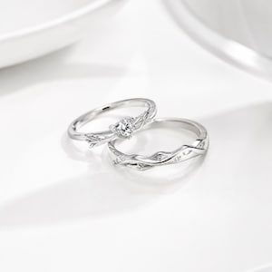 May include: Two silver-toned rings with a polished finish. One ring features a single round clear stone set in a band with a textured, branch-like design. The second ring is a matching band with a continuous, intertwined vine or branch pattern.
