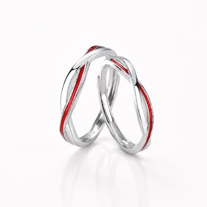 Può includere: Due anelli color argento con finitura metallica lucida e un design a fascia intrecciata continua. Ogni anello è accentuato da un sottile inserto rosso vibrante che segue la curva del metallo, creando un aspetto elegante e riflettente.