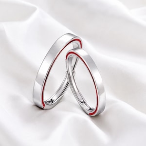 Könnte beinhalten: Zwei silberfarbene Ringe mit glatter, polierter Oberfläche und einer dünnen, leuchtend roten Linie, die mittig entlang des Rings verläuft. Die Ringe haben ein offenes, verstellbares Design, das eine Anpassung an verschiedene Fingerbreiten ermöglicht.