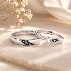 May include: Two silver-toned rings featuring a leaf motif with dark green enamel inlay. One ring is a simple band, while the other is accented with a small clear stone and a round, iridescent white stone, both designed with an open adjustable band.