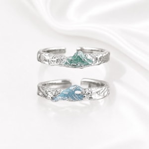 Può includere: Due anelli aperti color argento con una superficie della fascia testurizzata e irregolare. Un anello presenta un piccolo design a rilievo di una cima di montagna verde, mentre l'altro anello mostra un motivo a rilievo di un'onda blu. Design regolabile.
