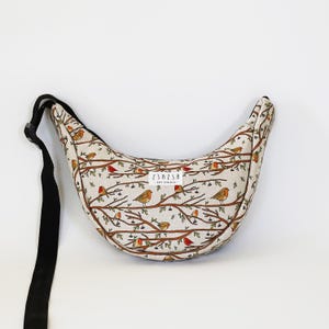 May include: A crescent-shaped crossbody bag in a light beige fabric featuring a repeating pattern of small orange and brown birds perched on brown branches. The bag includes a black adjustable strap and a white label reading ZSAZSA ART STUDIO.