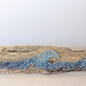 May include: A string art piece on a long piece of driftwood featuring a yellow sun with rays and blue ocean waves created with nails and thread. The design transitions from dark blue to light blue across the length of the textured wooden base.