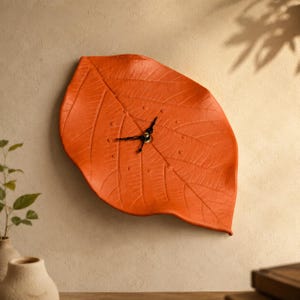 Pode incluir: Um relógio de parede em forma de uma folha laranja grande e texturizada. O mostrador apresenta uma nervura central proeminente e nervuras menores que se ramificam pela superfície, com ponteiros pretos de horas e minutos no centro.
