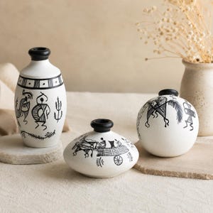 Peut inclure: Trois vases en céramique blanche avec des bords noirs et des illustrations au trait noir peintes à la main. La collection comprend un vase cylindrique haut, un vase large et bas, et un vase sphérique, chacun orné de figures tribales.