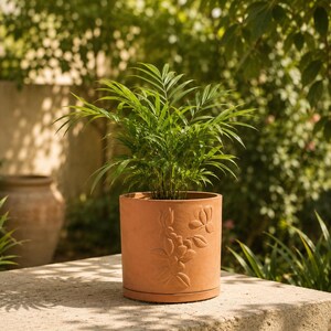 以下が含まれることがあります： 前面に花のレリーフが施された円筒形のテラコッタ製植木鉢。羽のような葉を持つ鮮やかな緑色のテーブルヤシが植えられています。鉢は同素材の円形受け皿の上に置かれており、自然な粘土の質感が特徴です。
