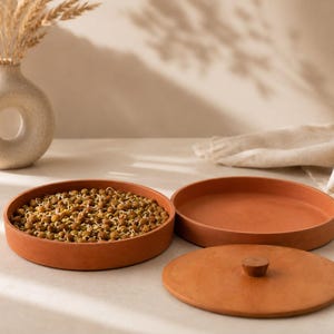 May include: Two round terracotta trays with low walls and a matching circular lid with a small central knob. One tray is filled with sprouted green legumes. The items are made of a matte, reddish-brown clay material with a smooth, uniform finish.