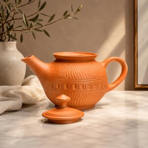May include: A terracotta teapot with a rounded body, a curved handle, and a short spout. The exterior features horizontal bands of textured, carved patterns. A matching circular lid with a small knob handle rests in front of the teapot.