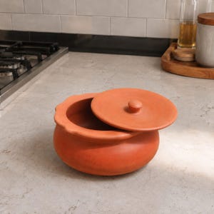 Puede incluir: Una olla de barro de color terracota con un acabado liso y mate y un borde festoneado. La olla cuenta con una tapa circular a juego con un pequeño pomo central, que descansa parcialmente abierta sobre el borde superior del recipiente.
