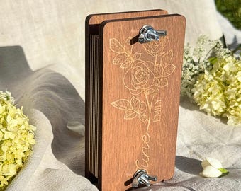 May include: A rectangular wooden flower press featuring two metal wing nuts for tightening. The front panel is engraved with a detailed rose illustration and the text "June" and "Emily" in a script font. The press consists of stacked wooden layers.