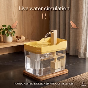 Op de afbeelding: Een rechthoekige drinkfontein voor huisdieren met een doorzichtige basis en een gele bovenkant. Een kleine tuit creëert een waterstraal. De tekst "Live water circulation" en "HANDCRAFTED & DESIGNED FOR CAT WELLNESS" is zichtbaar.