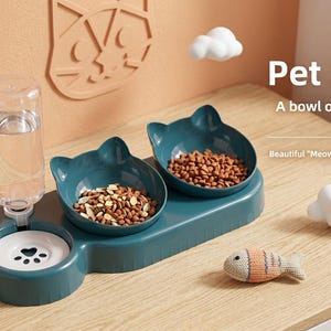 Puede incluir: Una estación de alimentación para mascotas de color verde azulado oscuro con dos cuencos en forma de orejas de gato llenos de comida seca y un recipiente de agua blanco con una huella. Una botella de agua de plástico transparente está unida al lateral. El texto dice Pet 3 bowl, A bowl of food and water snacks is done, Beautiful Meow Sheng has food and drink, y SHOW.