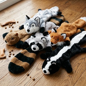 Könnte beinhalten: Eine Sammlung von fünf Plüschtier-Spielzeugen, darunter ein grauer Wolf, ein braunes Eichhörnchen, ein grauer Waschbär mit gestreiftem Schwanz, ein rotbrauner Fuchs und ein schwarz-weißes Stinktier, verstreut mit kleinen beigen Leckerlis auf einem Holzboden.