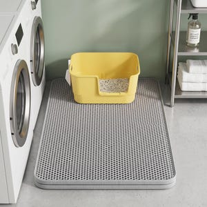 May include: A rectangular grey cat litter mat with a honeycomb-patterned top surface, positioned on the floor. A yellow plastic cat litter box with a low front entry and light-coloured litter inside sits directly on top of the grey mat.