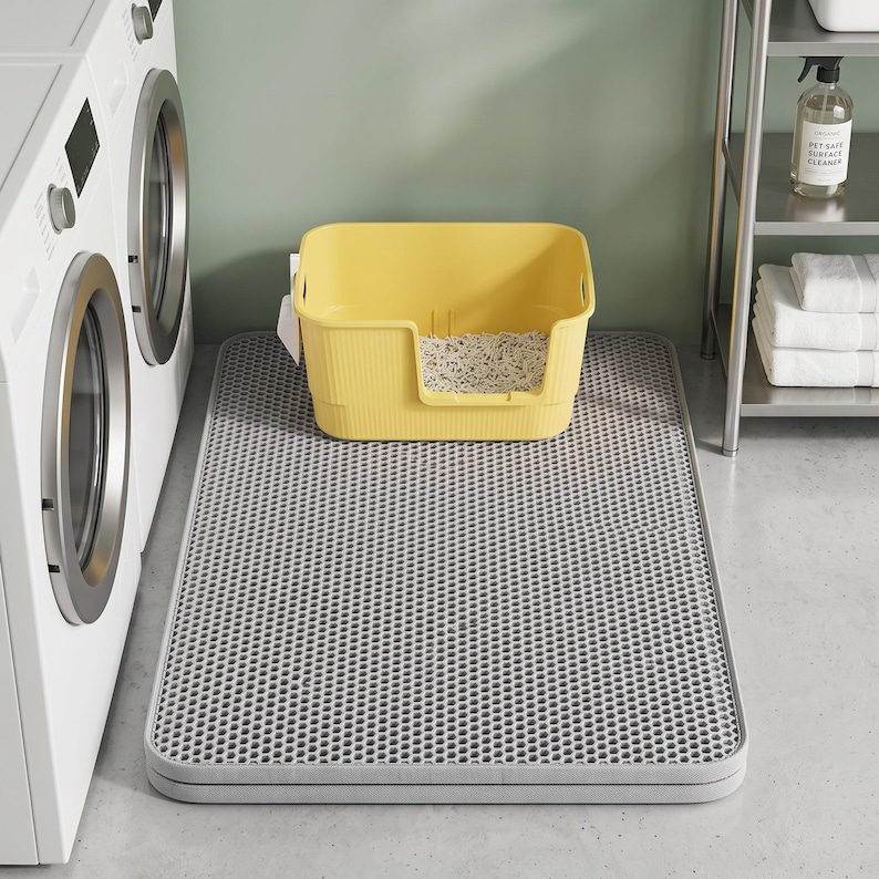 May include: A rectangular grey cat litter mat with a honeycomb-patterned top surface, positioned on the floor. A yellow plastic cat litter box with a low front entry and light-coloured litter inside sits directly on top of the grey mat.