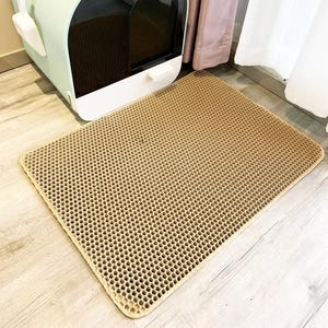 May include: A rectangular tan cat litter mat featuring a honeycomb-patterned top surface with numerous small hexagonal holes designed to trap debris. The mat is positioned on a light-coloured floor in front of a mint green and white enclosed cat litter box.