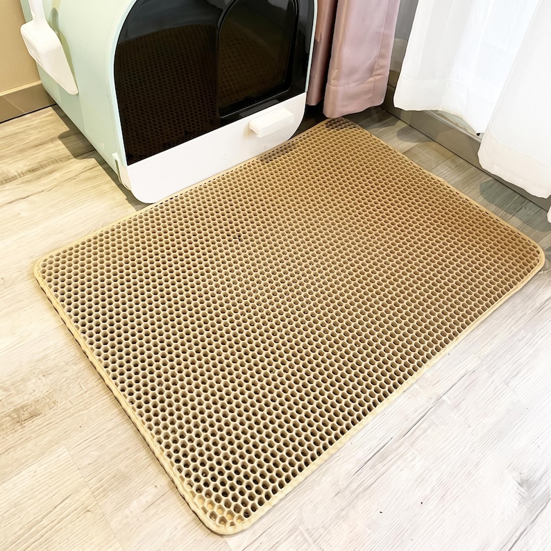 May include: A rectangular tan cat litter mat featuring a honeycomb-patterned top surface with numerous small hexagonal holes designed to trap debris. The mat is positioned on a light-coloured floor in front of a mint green and white enclosed cat litter box.
