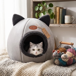 Pode incluir: Uma cama para animais de estimação cinza-claro em forma de cúpula com orelhas de gato cinza-escuro e uma abertura circular. Um gato fofo está dentro. Uma etiqueta castanha diz "HANDMADE WITH LOVE - COZY CAT CRAFTS". Cesto com novelos e ratos.