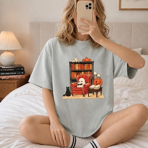 Könnte beinhalten: Ein hellgraues T-Shirt mit einem Grafikdruck, der einen Geist zeigt, der in einem roten Sessel sitzt und ein Buch liest. Die Szene umfasst eine schwarze Katze, ein Bücherregal voller Bücher, einen Kürbis, zwei Totenköpfe und einen kleinen Tisch.