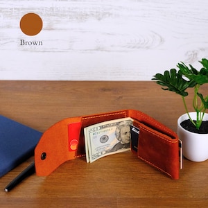 May include: A brown leather bifold wallet featuring visible stitching, a snap button closure, and internal slots containing a twenty-dollar bill and multiple credit cards. The wallet is open, displaying its interior compartments and contents.