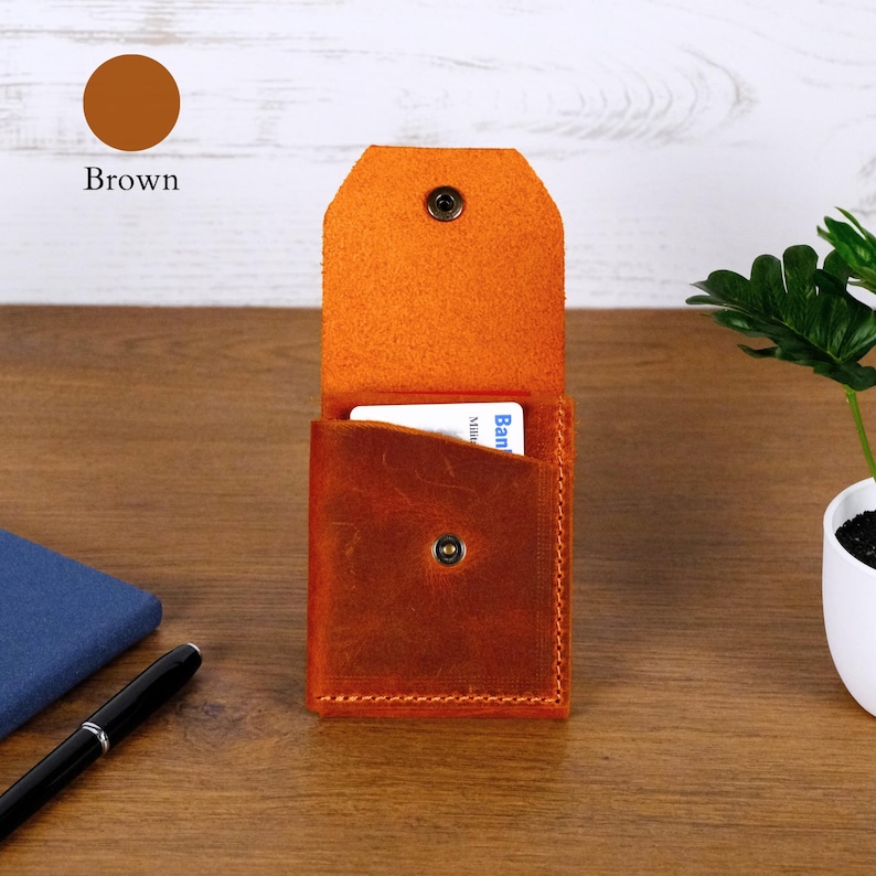 May include: A brown leather card holder with a vertical orientation, featuring a fold-over flap with a metal snap button closure. The interior pocket contains a white card with visible text. The leather has a textured, matte finish with visible stitching along the edges.