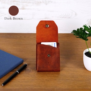 May include: A dark brown leather card holder with a vertical flap and a metal snap button closure. The wallet features visible stitching along the edges and contains a white card with blue text partially inserted into the main compartment.