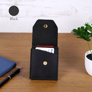 May include: A black leather card holder with a vertical orientation, featuring a fold-over flap secured by a brass-colored metal snap button. The front pocket contains a white card with visible text, and the exterior has a matte finish.