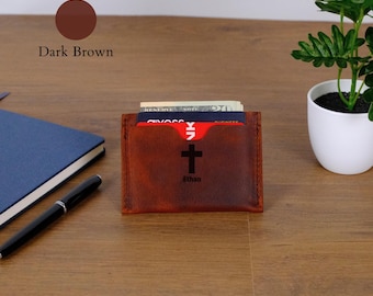 May include: A dark brown leather card holder featuring a black cross symbol and the name Ethan engraved on the front. The card holder contains several cards and a twenty-dollar bill, with visible stitching along the edges of the leather material.