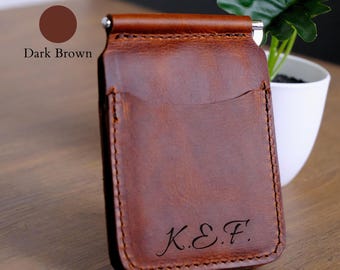 May include: A dark brown leather money clip wallet featuring a metal spring-loaded clip at the top and a curved card slot pocket on the front. The lower portion of the wallet is engraved with the stylized capital letters K.E.F. in a script font.