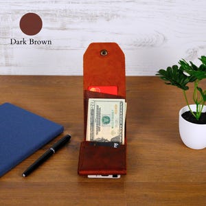 May include: A dark brown leather vertical wallet with a snap button closure, containing a twenty-dollar bill and multiple credit cards. The wallet features a tall, rectangular design with visible stitching along the edges and a flap top.