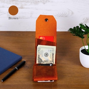 May include: A brown leather vertical wallet featuring a snap button closure, multiple interior slots for cards, and a central compartment containing a twenty-dollar bill. The wallet has a textured finish and a tall, rectangular structure.