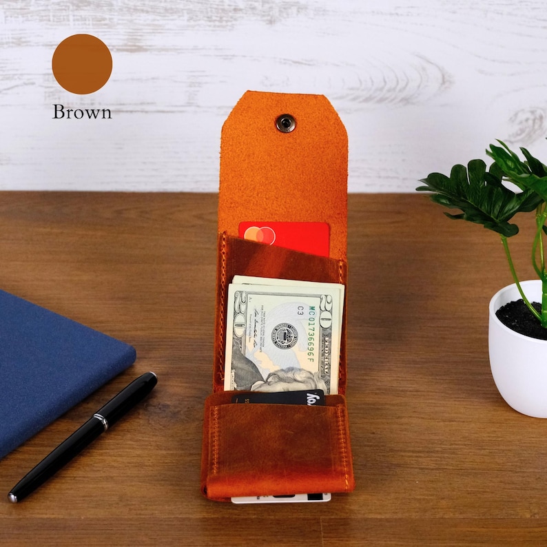 May include: A brown leather vertical wallet featuring a snap button closure, multiple interior slots for cards, and a central compartment containing a twenty-dollar bill. The wallet has a textured finish and a tall, rectangular structure.