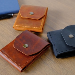 May include: Three small leather wallets with fold-over flaps and metal snap closures. One wallet is light tan, one is a deep reddish-brown, and one is black. Each wallet features visible stitching along the edges and a smooth, matte finish.