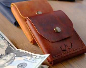 May include: Two leather wallets with snap closures, one tan and one dark brown. The dark brown wallet features an engraved letter E flanked by two antlers. A stack of twenty-dollar bills is visible in the foreground, partially obscuring the view.