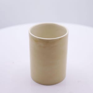 May include: A cylindrical ceramic cup with a smooth, light beige finish. The vessel features a simple, open-top design with subtle, soft tonal variations across its exterior surface, creating a clean and uniform appearance for this drinking container.