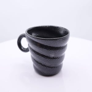 May include: A black mug with a textured, bumpy surface and a distinct spiral, ribbed pattern wrapping around the body. The mug features a small, circular handle attached to the side, and the overall finish is dark, glossy, and uniform in color.