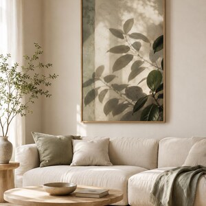 May include: A rectangular wall art print in a light wood frame featuring a soft-focus photograph of dark green leaves casting shadows against a textured beige wall. The artwork displays a play of light and shadow with muted earth tones.