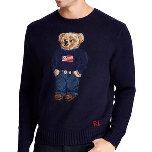 Könnte beinhalten: Ein dunkelblauer Langarmpullover mit Rundhalsausschnitt und einem mittigen Intarsien-Strickmotiv eines Teddybären, der einen blauen Pullover mit amerikanischer Flagge, eine blaue Jeans und braune Schuhe trägt. Die Buchstaben RL sind unten links rot gestickt.