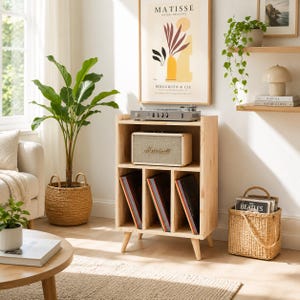 May include: A light-colored wooden record storage cabinet with four tapered legs. The top shelf holds a silver turntable and a beige speaker. The bottom section features three vertical dividers containing vinyl records. The unit is made of wood.