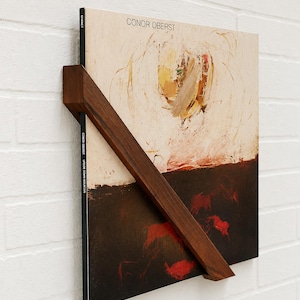 May include: A dark brown wooden wall-mounted vinyl record display shelf holding a Conor Oberst Upside Down Mountain album. The record cover features an abstract painting with a cream upper section and a dark brown lower section with red animal figures.